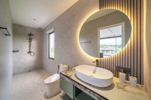 A modern bathroom with a large oval mirror, a round sink, a toilet, and a shower. - Villa Velora (Ubud)