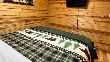 Una habitación de cabaña de madera con una cama, un cuadro de un paisaje forestal y un televisor montado.