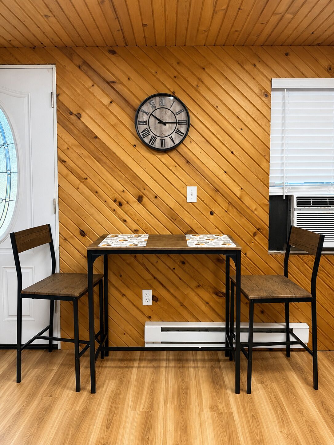 Una habitación con paneles de madera, un reloj sujeto a la pared, una mesa con sillas y un equipo de aire acondicionado.