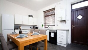 Een kleine keuken met witte kastjes, een houten eettafel en een donkerhouten deur.