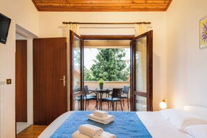 Ein Schlafzimmer mit Bett, Fenster, Tür, einem Tisch mit Stühlen und einer Zimmerpflanze vor dem Fenster.