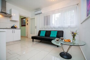 A modern kitchen with white cabinets, a black sofa with teal cushions, a glass table with a cup and croissants, and a window with sheer curtains. - Marisa Olive Grove - App Luna, Labin, Croatia (Labin)