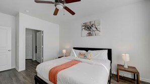 Un dormitorio con cama, ventilador de techo, mesita de noche con lámpara y un cuadro en la pared.
