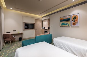 Habitación de hotel con dos camas individuales, un escritorio, una silla, un televisor y obras de arte en la pared.