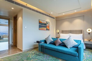 Una habitación de hotel moderna con un sofá color turquesa, una cama con ropa blanca, una mesita de noche pequeña y una lámpara de pared.