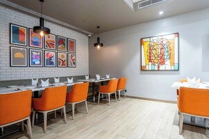 Un restaurante con una decoración moderna, que presenta una pared de ladrillo con obras de arte enmarcadas, sillas anaranjadas y un amplio ventanal de vitral.