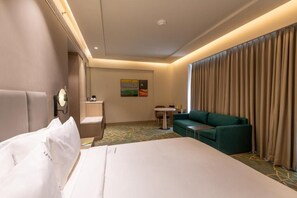 A hotel room with a bed, a green sofa, a small table, a chair, and a painting on the wall. - Holiday Inn Katra Vaishno Devi (Reasi)