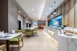 A modern hotel dining area with long tables, green chairs, a buffet counter, and a television screen.