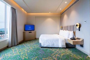 A hotel room with a bed, a television on a stand, a nightstand, and a window with curtains.
