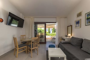 Ein Wohnzimmer mit Sofa, einem Wand-Fernseher, einem Esstisch mit Stühlen und einem Balkon mit Blick auf Grünflächen.