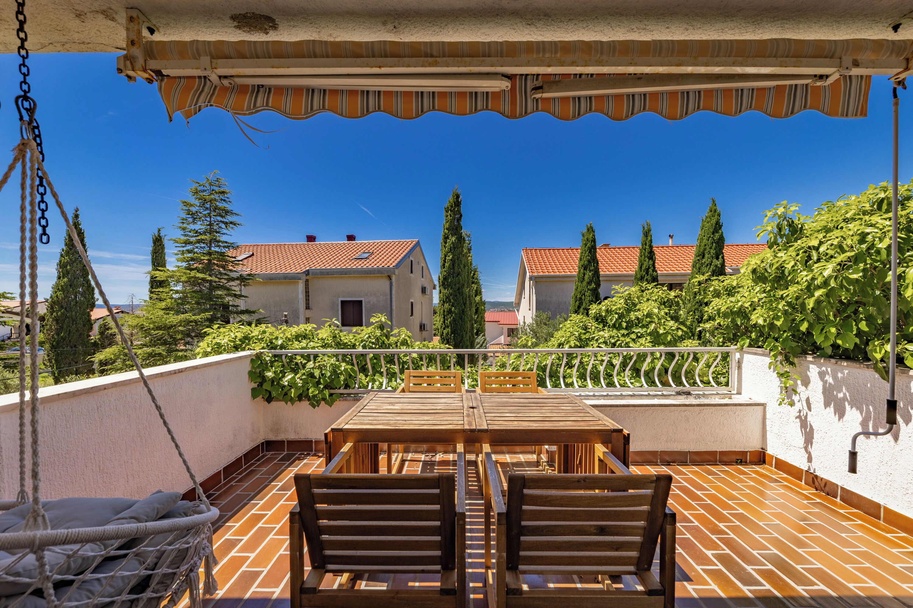 Terraço no telhado com mesa e cadeiras de madeira, uma cadeira suspensa e vista para casas e árvores.