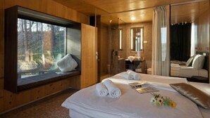 Quarto estilo cabana de madeira com uma janela grande, cama com toalhas dobradas, banheiro com espelho e um espaço de estar.