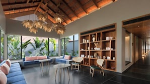 A modern interior with a high ceiling, a large bookshelf, a seating area with sofas and chairs, and large windows with plants.