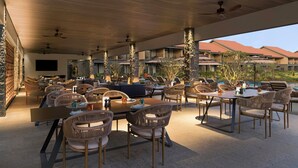 An outdoor dining area with multiple tables and chairs, ceiling fans, and a view of hotel buildings in the background.