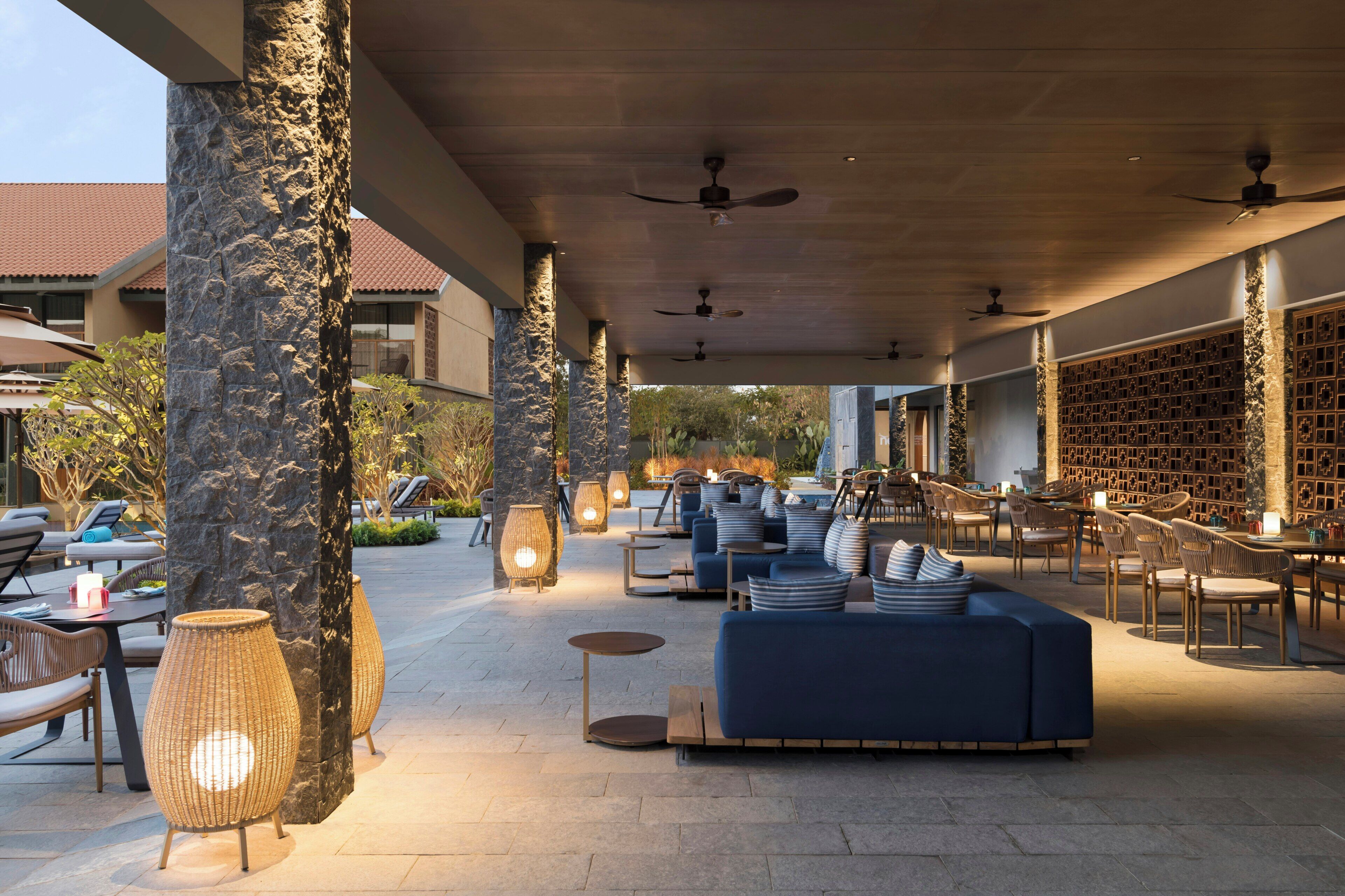 An outdoor seating area with a blue sofa, wicker chairs, and tables, surrounded by stone pillars and a ceiling fan.