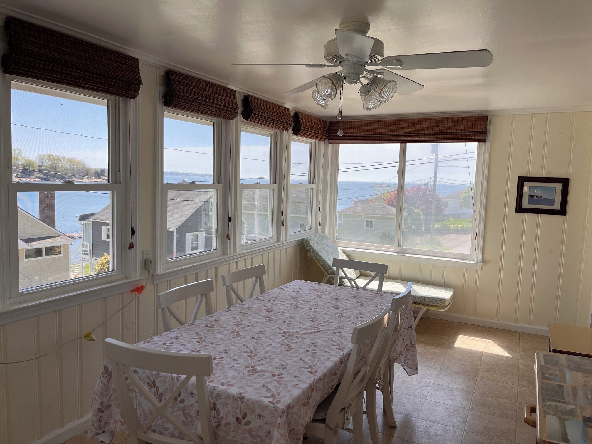Une pièce avec une table et des chaises, un ventilateur de plafond, de grandes fenêtres équipées de stores et une vue sur la mer.