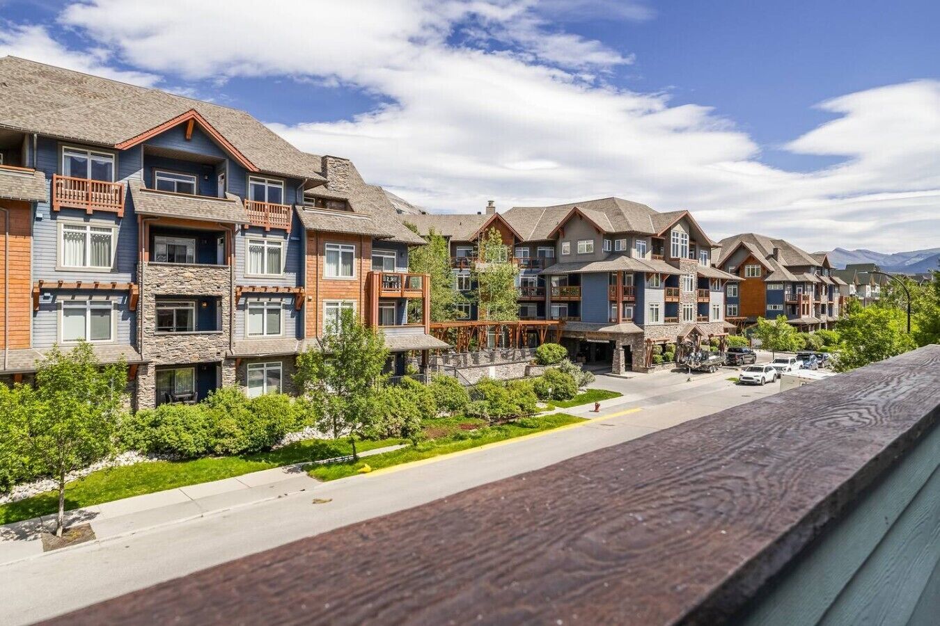 A row of multi-story buildings with balconies, a paved street, and greenery.