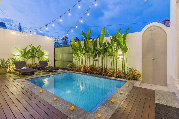 A swimming pool with a wooden deck, lounge chairs, and string lights. - Vinyak Villa (Kerobokan)
