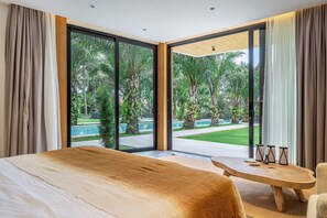 Un dormitorio con una cama grande, una mesita de noche de madera y vistas a una piscina y vegetación a través de puertas correderas amplias.