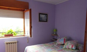 Un dormitorio con cama, ventana con persianas, radiador, cuadro enmarco, mesita de noche con lámpara y planta en maceta.