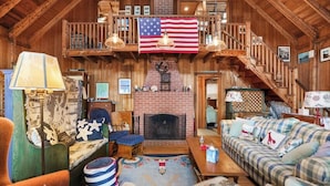 A rustic interior with a fireplace, American flag, wooden furniture, and a staircase. - Charming 4BR, 2BA Southport retreat for up to 8 guests. (Southport)