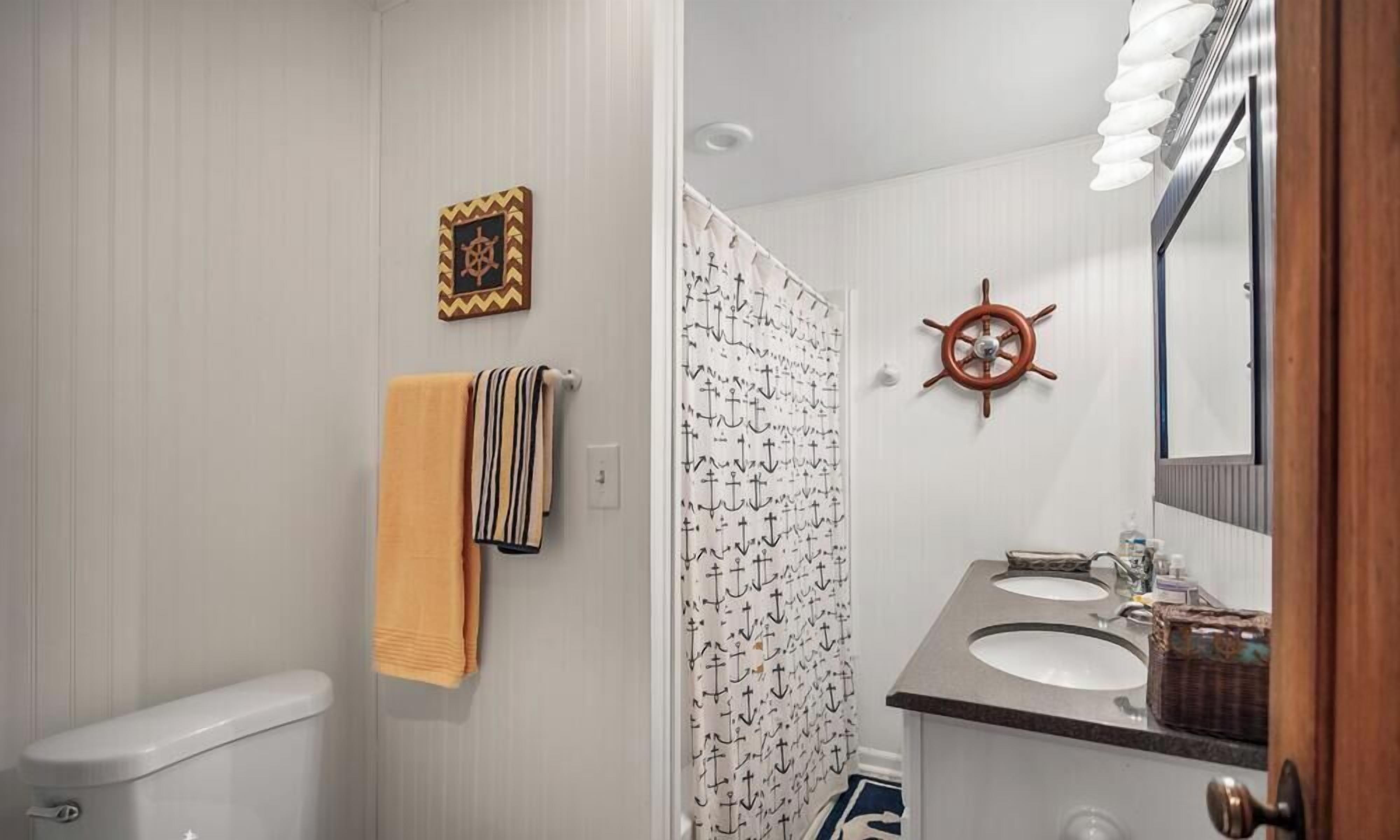 A bathroom with a toilet, a towel rack with a striped towel, a ship's wheel decoration, and a shower curtain with anchor patterns.