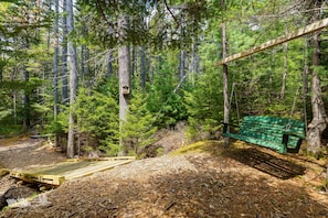 A forested area with a wooden deck, a green bench, and a birdhouse. - Waterfront 3BR East Boothbay retreat with ocean views, deck, WiFi. (East Boothbay)