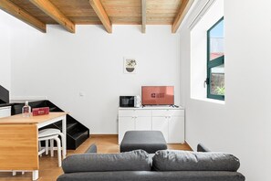 A modern, compact living space with a wooden ceiling, a desk with a computer and a TV, a grey sofa, and a window with a view. - GuestReady - Trendy House (Porto)