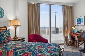 A hotel room with a bed, a desk, a chair, a lamp, and a view of the beach through the balcony doors. - Sea Breeze Beachfront Walkout Poolside Condo (Biloxi)