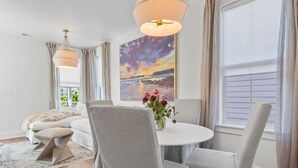 Ein modernes Wohnzimmer mit einem weißen Sofa, grauen Sesseln, einem runden Esstisch und einem Gemälde, das einen Sonnenuntergang zeigt.