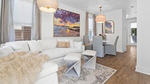 Ein modernes Wohnzimmer mit einem weißen Ecksofa, einem Essbereich mit grauen Stühlen und einem großen Gemälde, das einen Sonnenuntergang zeigt.