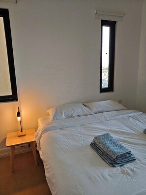 Ein Schlafzimmer mit Bett, Nachttisch, Lampe, Fenster und Blick ins Freie.
