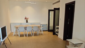 Quarto moderno e bem iluminado com mesa de madeira, sofá, televisão em suporte e área de jantar com cadeiras e um vaso de flores.