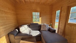 A wooden cabin interior with a gray sectional sofa, a small wooden table, and two windows offering a view of the outdoors. - The Pine Loft Bunkie (Lac du Bonnet)