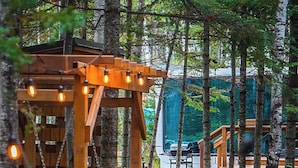 A wooden structure with a platform and steps, surrounded by trees and a vehicle in the background. - The Pine Loft Bunkie (Lac du Bonnet)