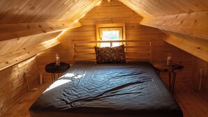 A loft-style bedroom with a wooden ceiling, a bed with a dark mattress, a window with a view, and a small table with a candle. - The Pine Loft Bunkie (Lac du Bonnet)