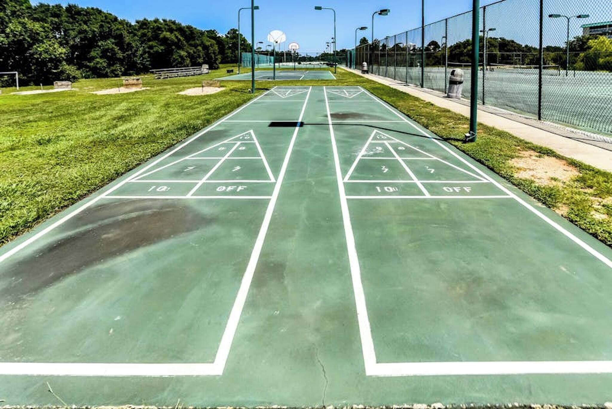Un terrain de jeu de shuffleboard avec des marquages verts et des lignes blanches, entouré d’une clôture et d’arbres.