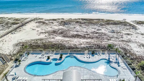 A pool with a wavy design, surrounded by lounge chairs and palm trees, overlooking a beach. - Beach View Plantation 4515 With Multiple Resort Amenities (Gulf Shores)