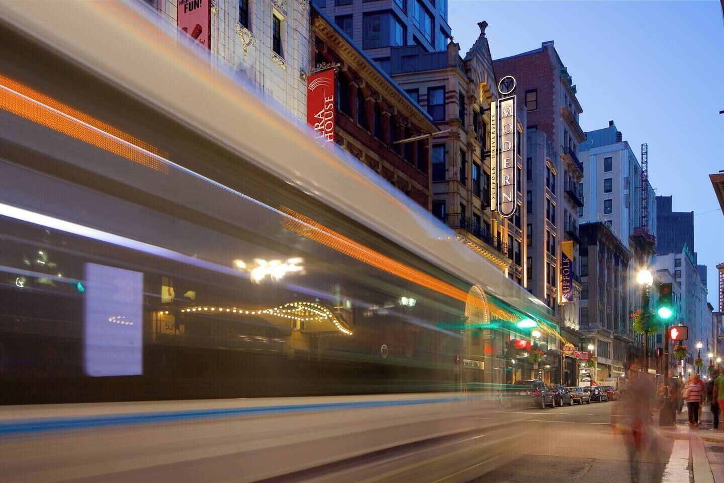 A train passing by a row of buildings with illuminated signs, including 'ERA HOUSE' and 'MODERN ZINC.'