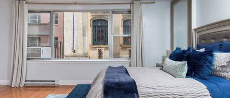 Un dormitorio con un ventanal grande, una cama con ropa de cama azul y blanca, un banco y vistas de un edificio.