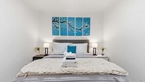 Una cama bien tendida con una manta blanca, una almohada azul y una toalla blanca encima. Hay dos mesitas de noche con lámparas y pequeñas plantas. Un cuadro grande de un árbol con flores está en la pared sobre la cama.