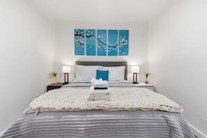 A neatly made bed with a white blanket, a blue pillow, and a white towel on top. There are two bedside tables with lamps and small plants. A large painting of a tree with blossoms is on the wall above the bed. - 7 Min Walk to Golden Belt | 8 Min to Hospital (Durham)