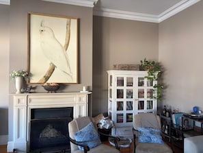A living room with a fireplace, a large framed artwork, a white cabinet with glass doors, and a small table with a chair. - Somewhere Special, Pool, Steam Sauna, Outdoor Bar, Fireplace (Woodville Park)