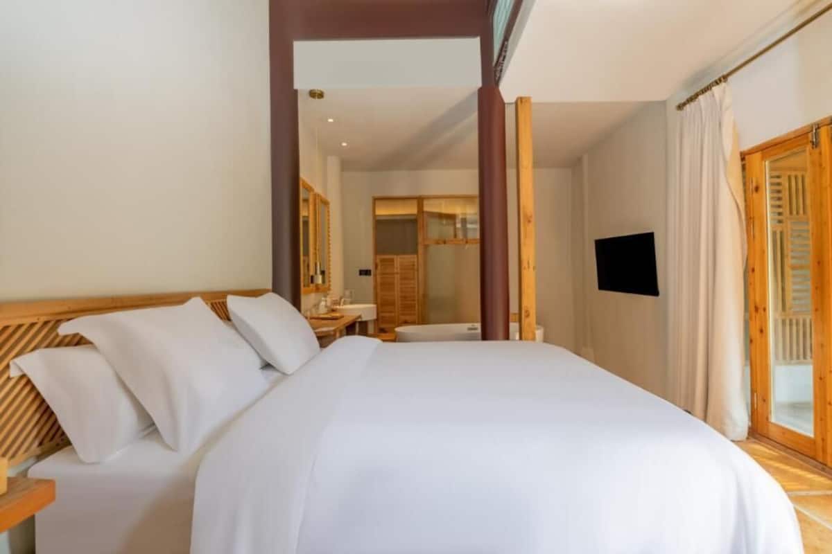 A hotel room with a bed, a TV, a wooden headboard, a bathroom visible through an open door, and a wooden floor.