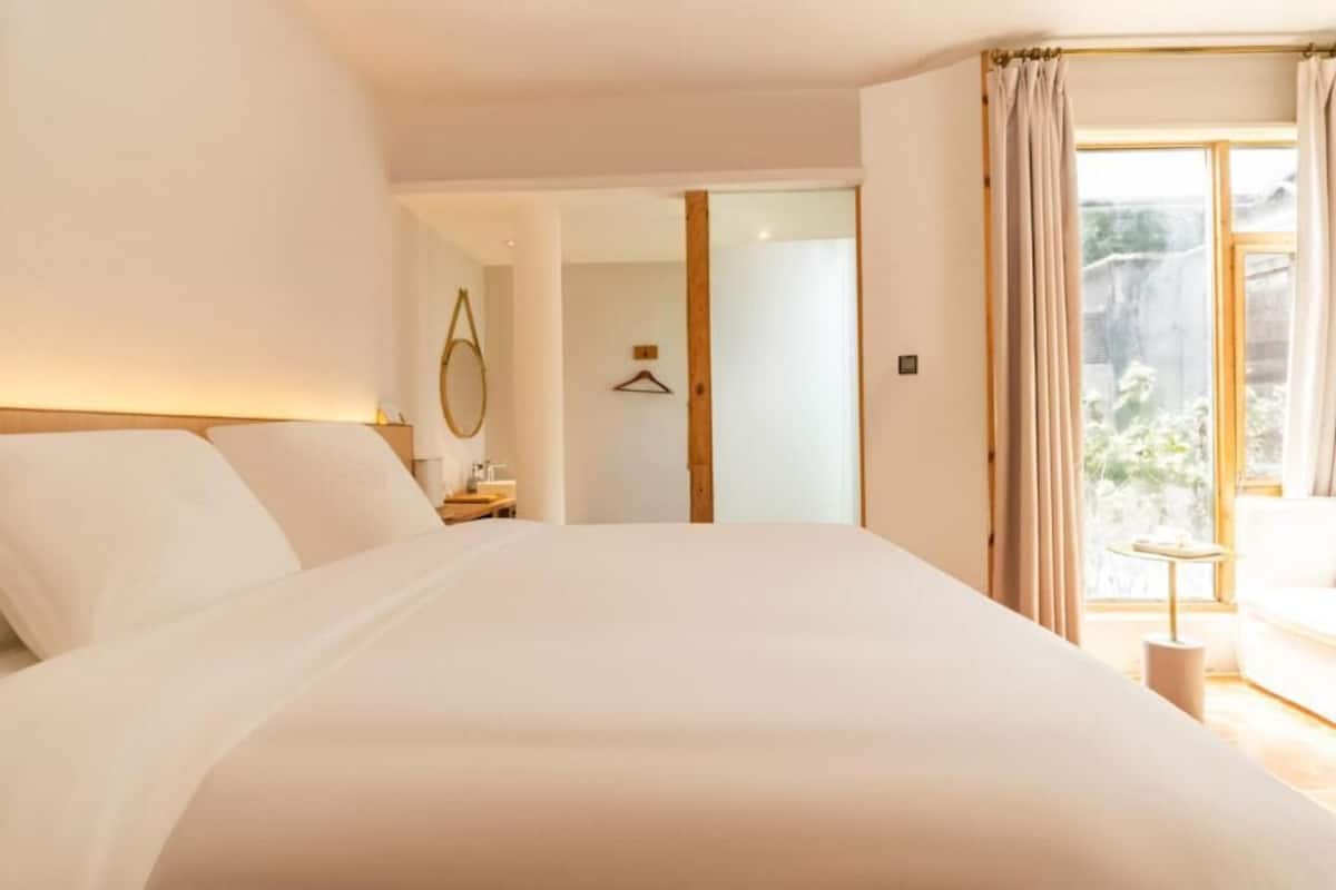 A bedroom with a large bed, a bedside table, a hanging light, and a view of the outdoors through a window.