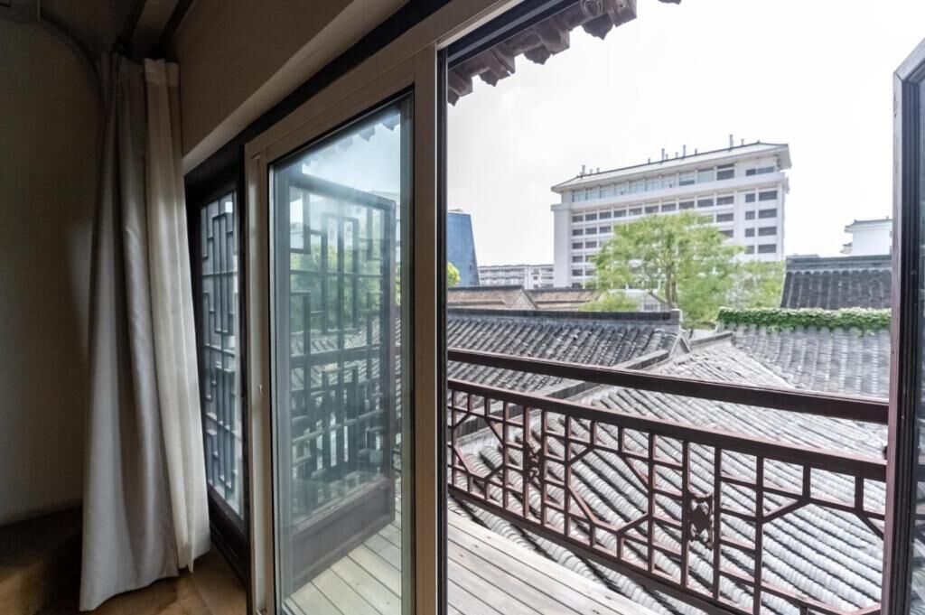 A room with a sliding glass door, a wooden balcony with a metal railing, and traditional tiled roofing visible through the window.
