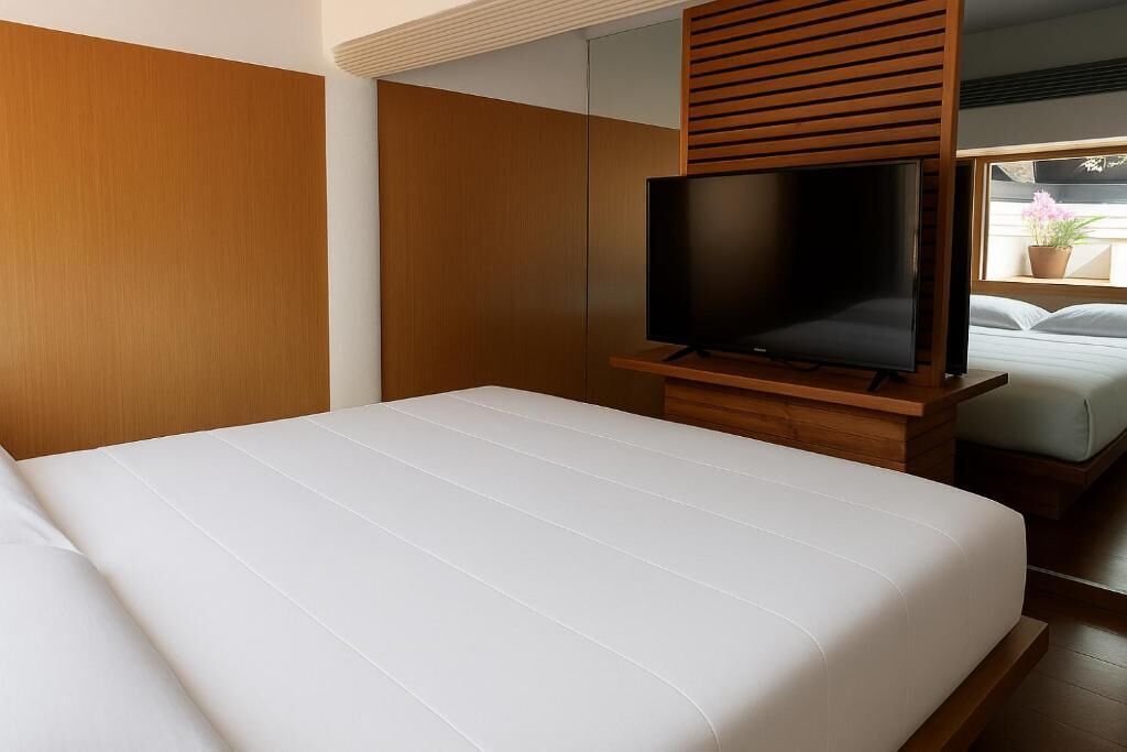 A hotel room with a bed, a TV on a wooden stand, and a window with a plant.