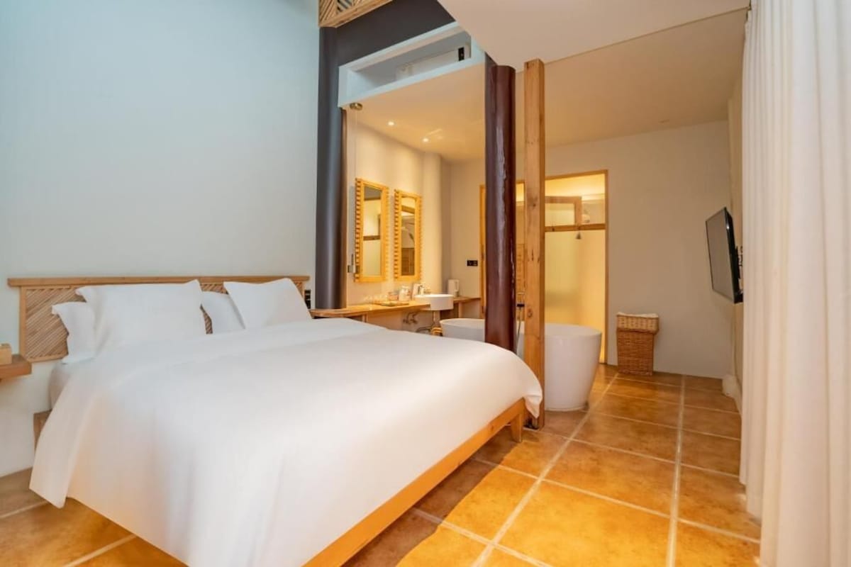 A hotel room with a large bed, a wooden headboard, a mirror, a bedside table, a TV, and a bathroom with a shower and a toilet.