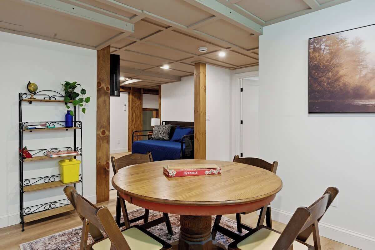 A room with a round wooden table, four chairs, a bookshelf, a bed with a blue blanket, and a painting on the wall.