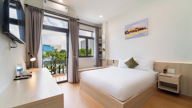 A modern hotel room with a large bed, a desk, a nightstand, and a balcony with a view of buildings and greenery. - SORA HOTEL & APARTMENT (Ho Chi Minh City)